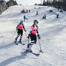 Zwei Skifahrerinnen mit pinken Leibchen fahren die Piste hinab. Die vorderer Skifahrerin fährt auf einem Ski und trägt auf dem Leibchen die Aufschrift Guide.